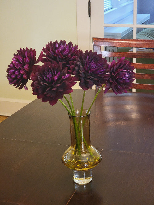Purple flowers in a clear vase on a reflective surface with a window in the background